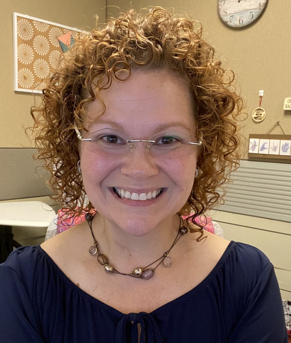 [ID: Headshot image of a white woman with curly red hair, glasses and a brass necklace glasses is pictured sitting in her office at the Center for Students with Disabilities (CSD)]