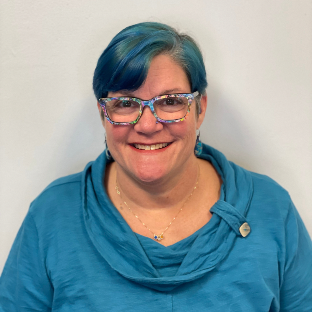 [ID: a white woman is pictured against a white wall wearing a teal blue sweater, teal blue glasses with teal colored short hair]